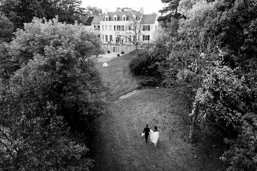 A couple walks through a wooded area back to a mansion at Harding Waterfront Estates. A wedding day drone photograph shot by Pixelesque Photography - Hamilton wedding Photographer.