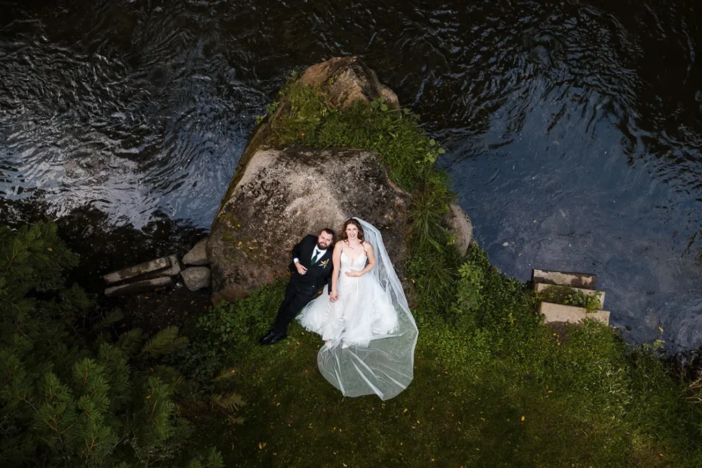 A wedding couple lay on a rock and look at the drone as a river rushes by them at Homegrown hideaway. A wedding day drone photograph shot by Pixelesque Photography - Hamilton wedding Photographer.