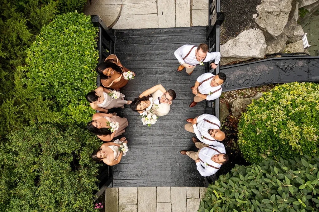 A couple and their surrounding wedding party stand on a bridge and look up at the drone at the Old Mill in Toronto. A wedding day drone photograph shot by Pixelesque Photography - Hamilton wedding Photographer.