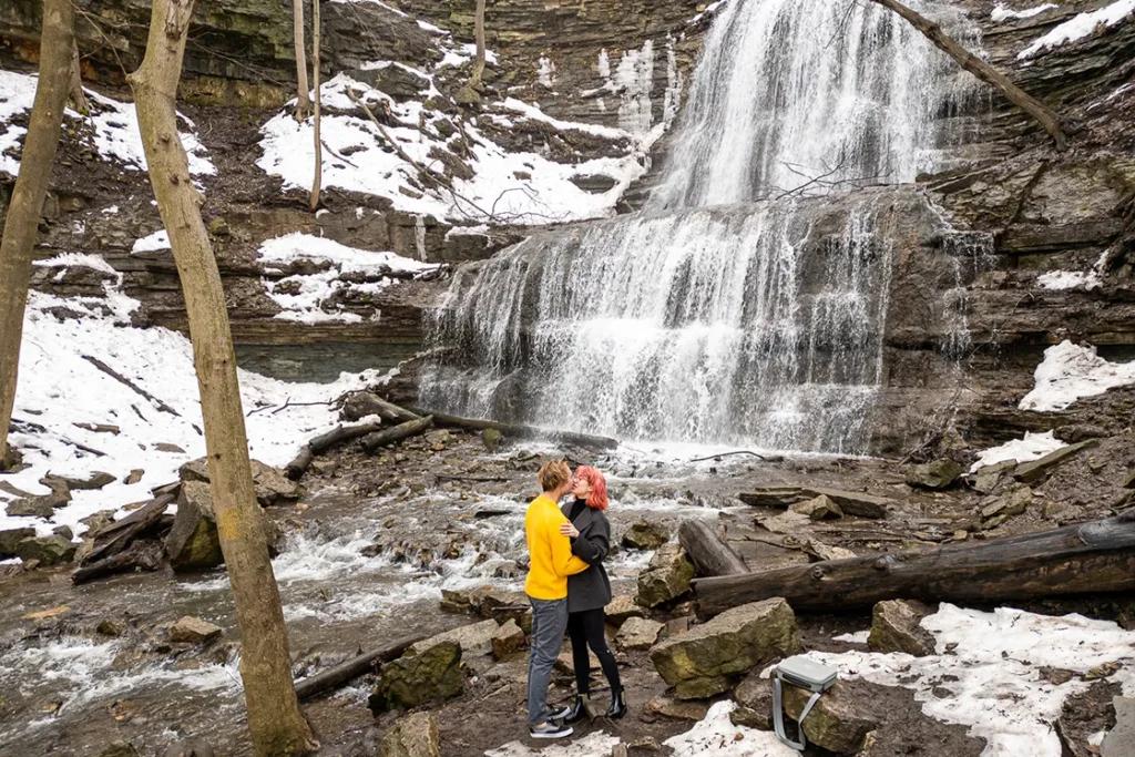 A couple shares a kiss as Sherman Falls and the snow covered rocks surround them. An engagement session drone photograph shot by Pixelesque Photography - Hamilton wedding Photographer.