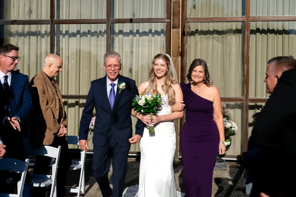 the bride walks arm in arm with her parents and laughs as she walks down the aisle.Shot at Carmen's event centre, Hamilton by Pixelesque Photography.