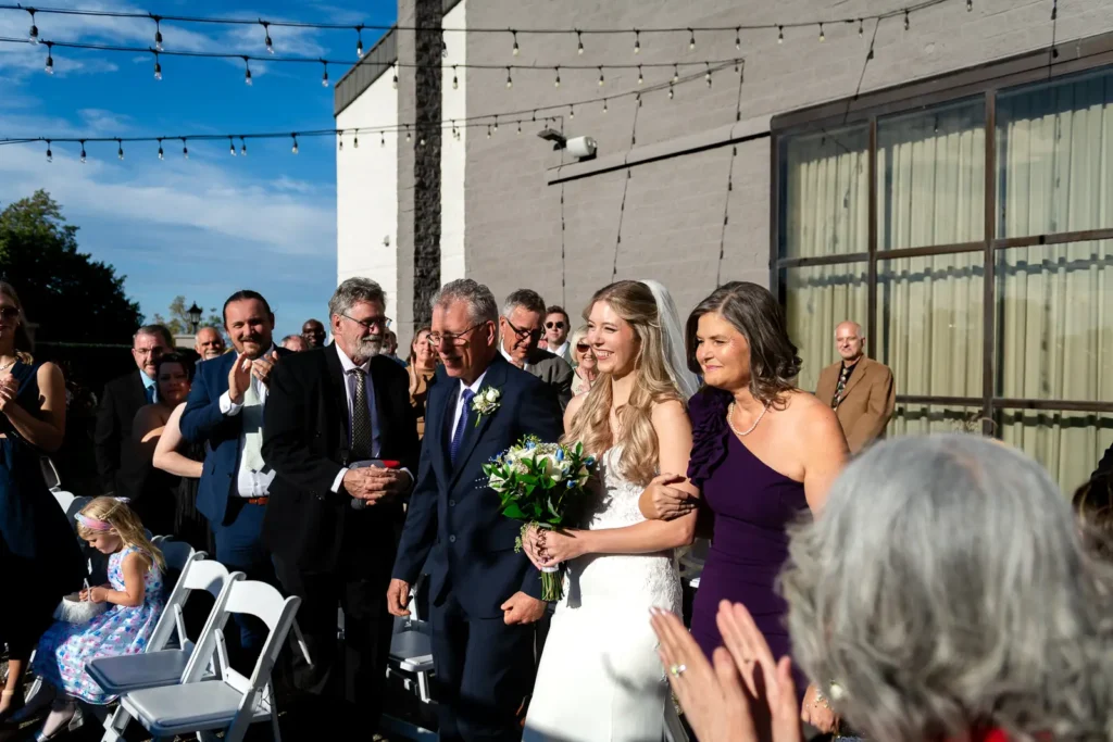 the bride laughs and people clap as she walks down the aisle with her parents. Shot at Carmen's event centre, Hamilton by Pixelesque Photography.