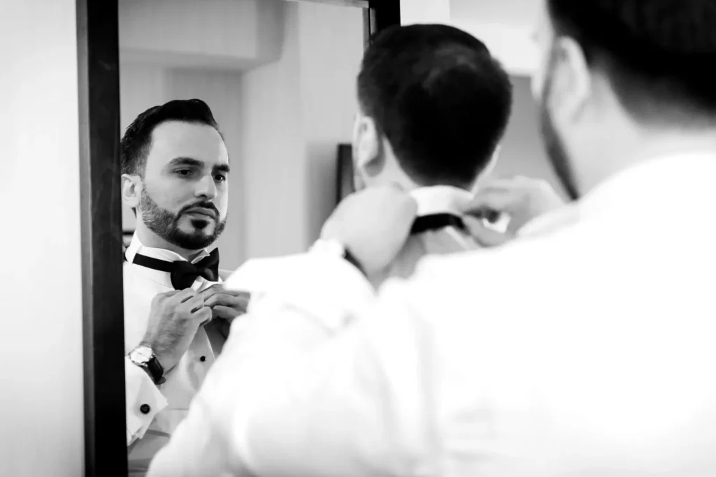 the best man helps the groom adjust his bow tie as the groom looks in the mirror. Shot in C hotel, Hamilton by Pixelesque Photography.