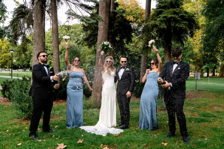 2 groomsmen pop champagne bottles as the couple and bridesmaids celebrate. Shot at Hamilton's Gage Park by Pixelesque Photography.