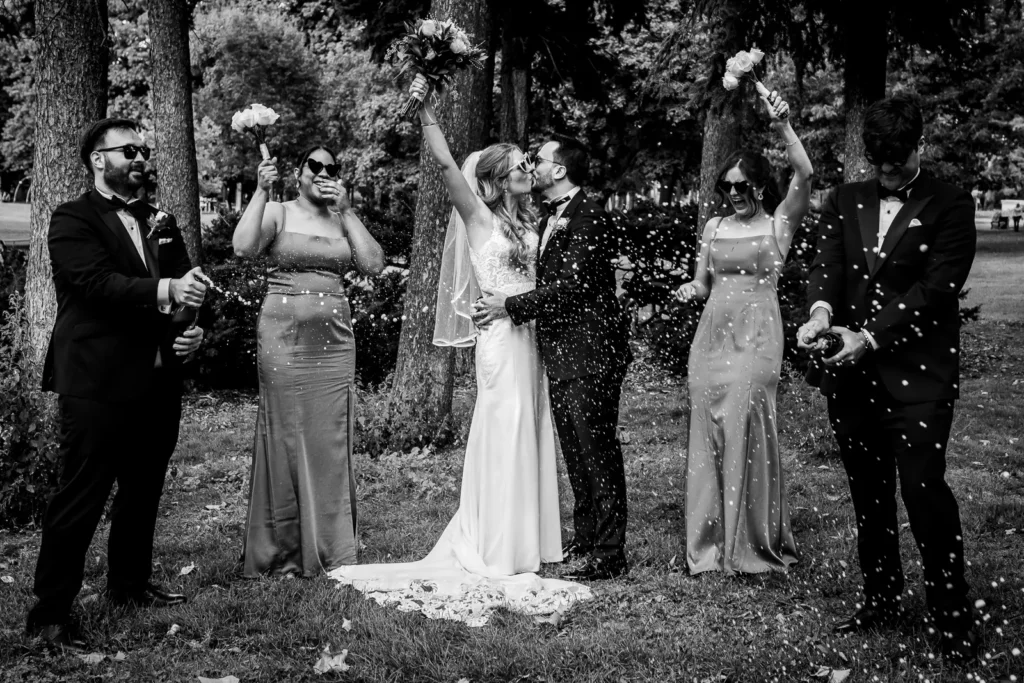 the wedding couple shares as a kiss in gage park as the bridal party pops champagne and dances around them. shot in hamilton by Pixelesque Photography.