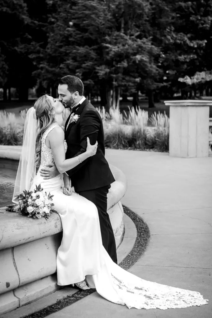 a groom kisses his bride who is sat on the edge of a stone fountain. Shot at Hamilton's Gage Park by Pixelesque Photography.