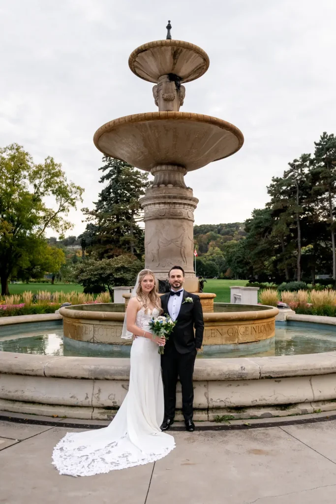 a wedding couple pose in front of a stone fountain. Shot at Hamilton's Gage Park by Pixelesque Photography.