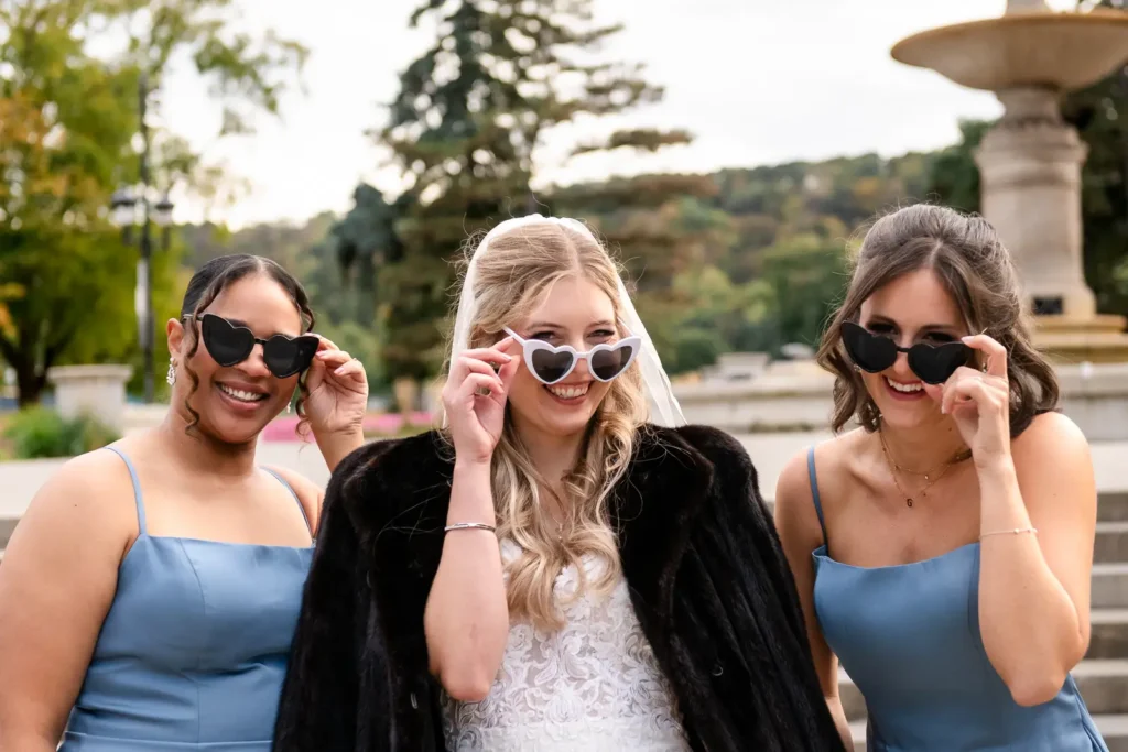a bride and her bridesmaids playfully lower their heart shaped sunglasses and look at the camera. Shot at Hamilton's Gage Park by Pixelesque Photography.
