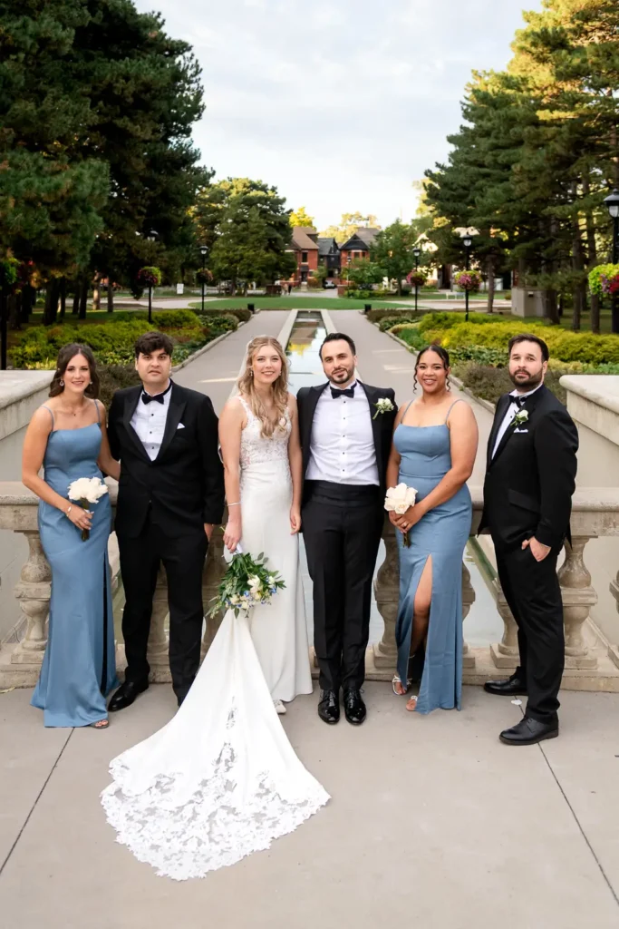 the bridal party stand in a line against a stone railing with a long reflecting pool behind them. Shot at Hamilton's Gage Park by Pixelesque Photography.