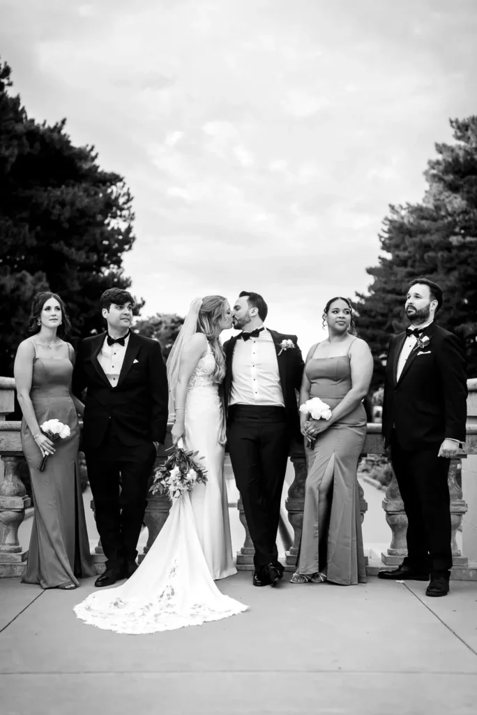 the wedding party stand in a line against a stone railing and the couple go in for a kiss. Shot at Hamilton's Gage Park by Pixelesque Photography.