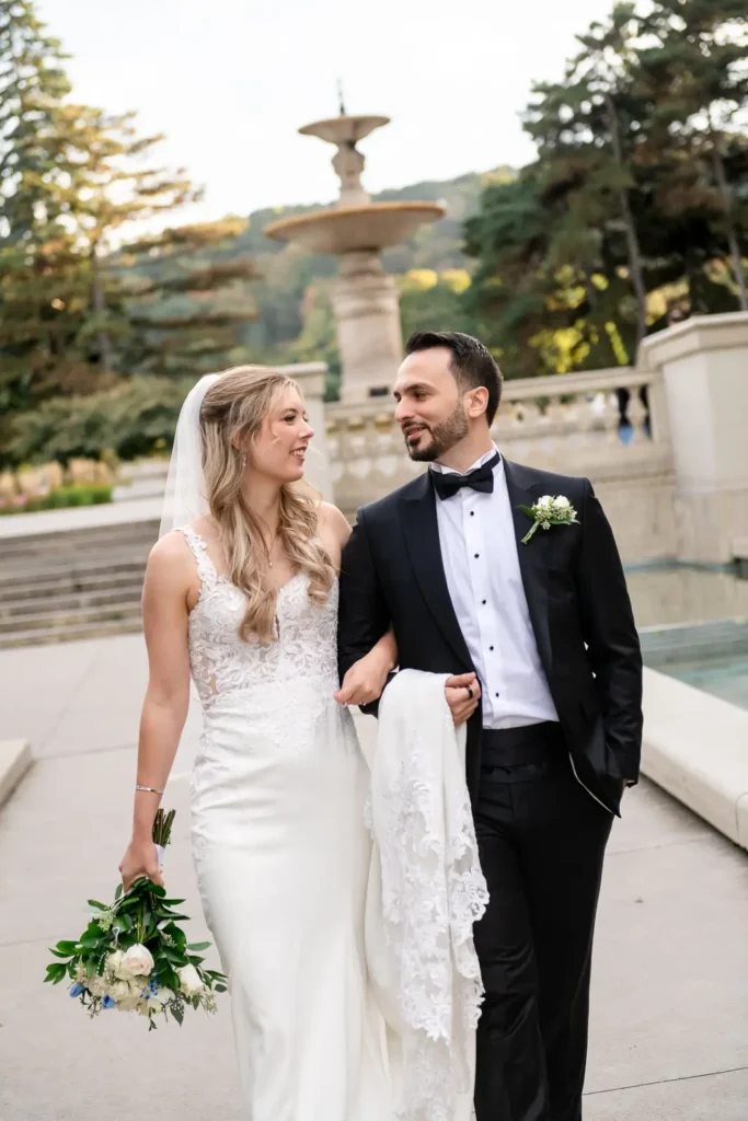 a groom walks along a fountain with his bride and holds her dress and she holds her bouquet. Shot at Hamilton's Gage Park by Pixelesque Photography.