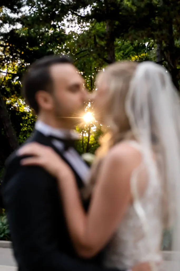 a wedding couple go for a kiss while they are out of focus and the sun peeking through the trees is in focus behind them. Shot at Hamilton's Gage Park by Pixelesque Photography.