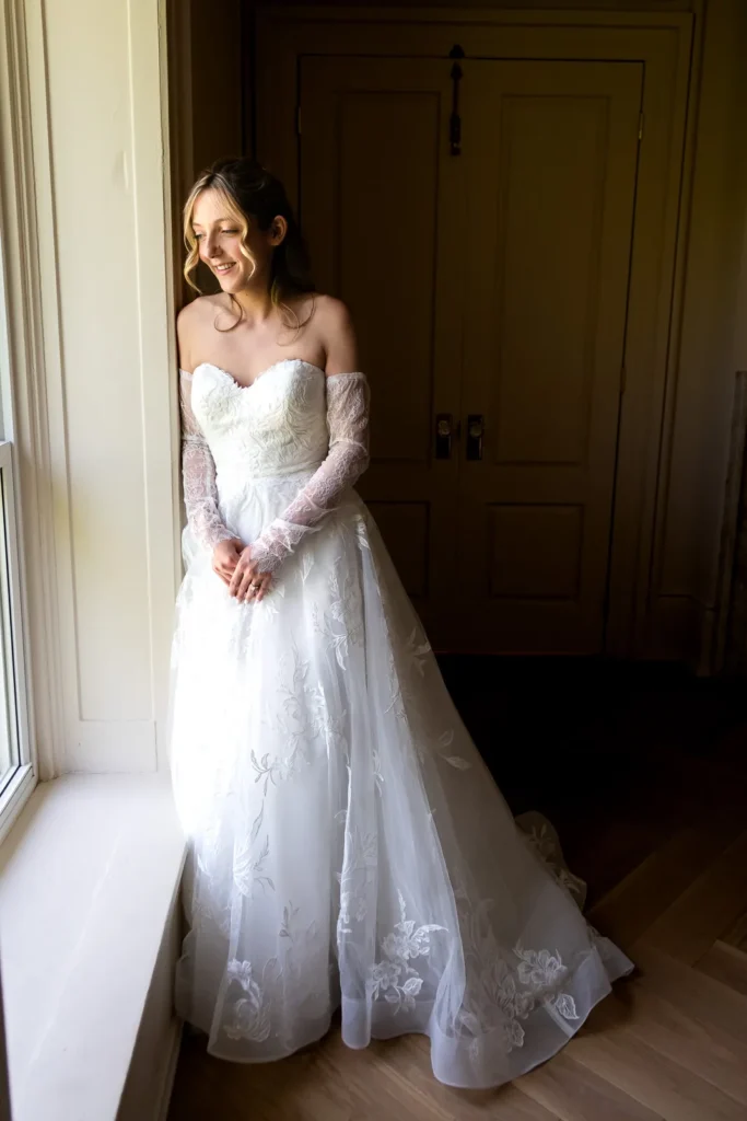Bride standing in her wedding dress beside a bright window at her family home.