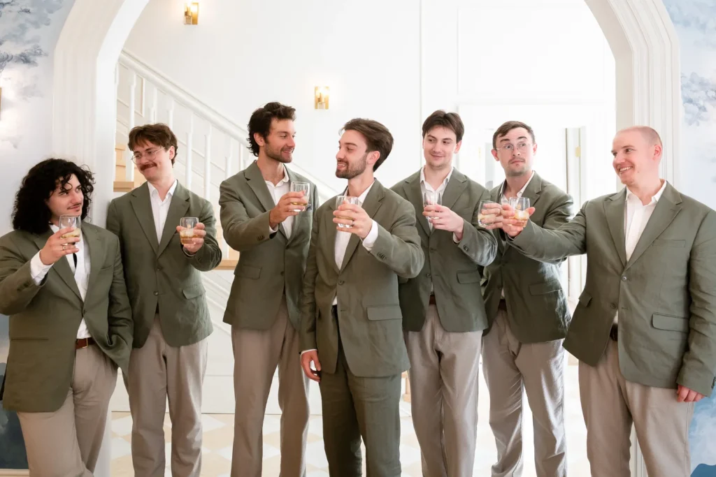 the groom and groomsmen share a drink indoors under an arch.