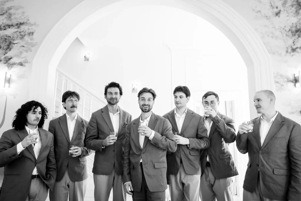 the groom and groomsmen share a drink indoors under an arch.