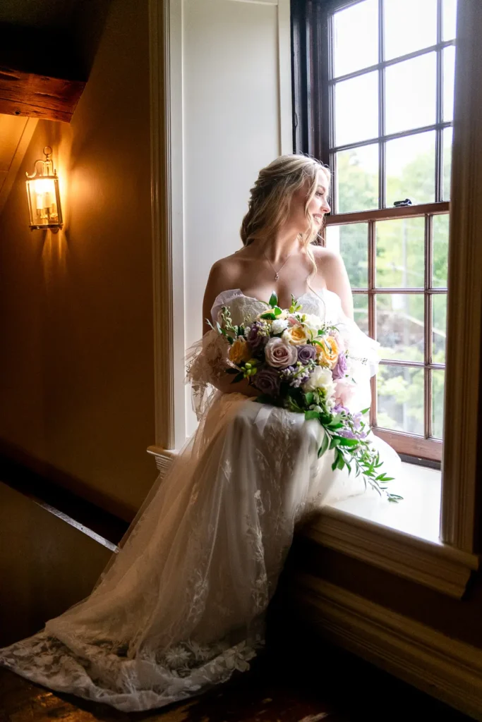 a bride sits on the window sill in her dress and with her bouquet at Ancaster Mill in Hamilton, Ontario.